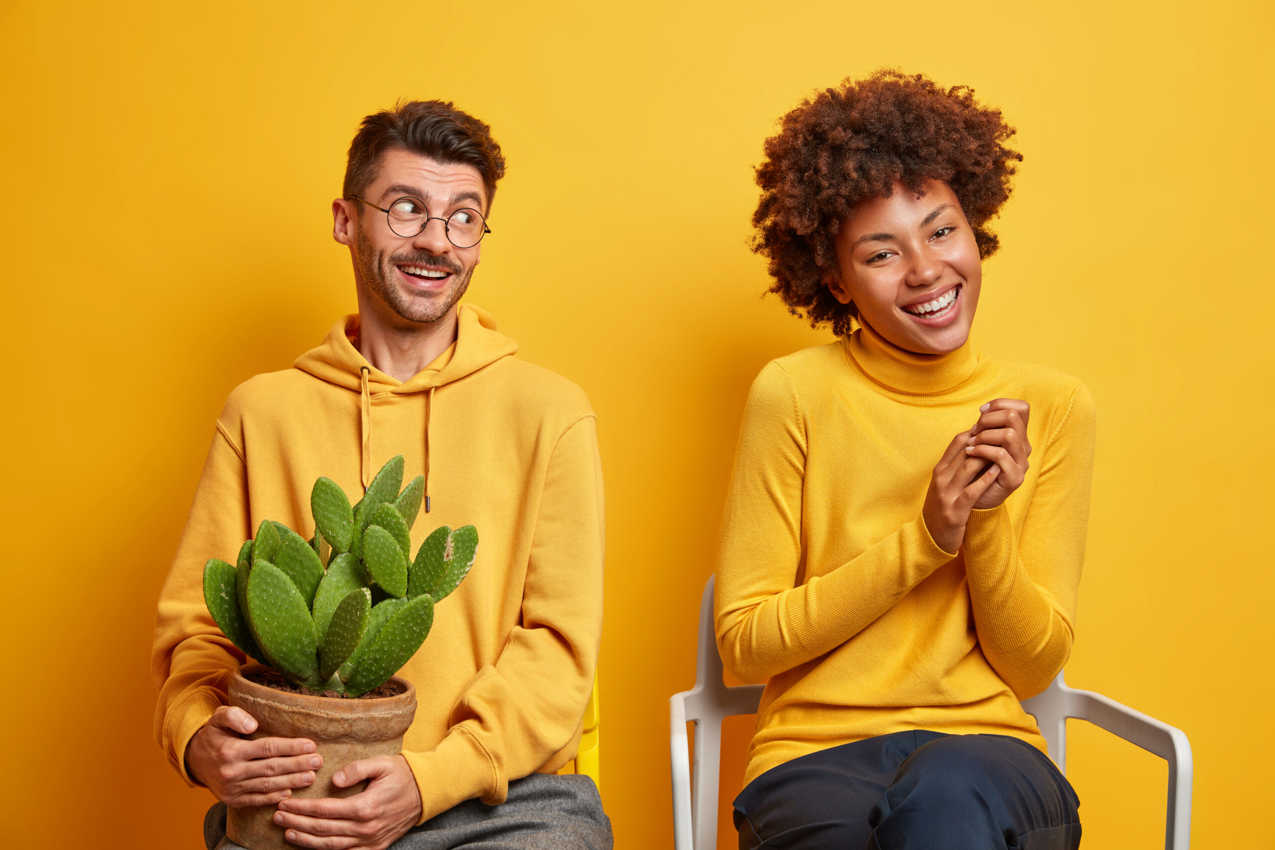 Home joyful mixed race woman and man laugh and have fun together pose on chairs against yellow background dressed casually express positive emotions spend free time at home. diverse family couple